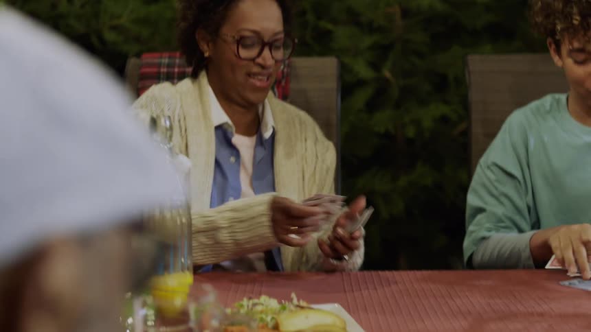 A mother plays a card game with her daughter and son as they all sit at an outdoor dining table with food and drinks at night beside a fireplace enjoying a good time together