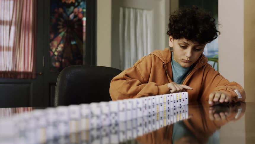 A boy concentrates while carefully arranging dominoes on a reflective table indoorsfocus tags
