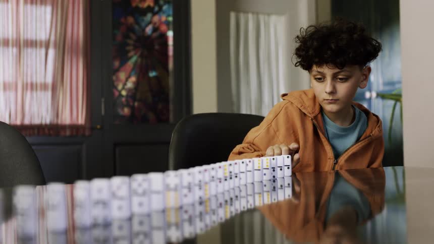 A boy in an orange jacket topples a long row of dominoes that stretches across the table toward the foreground