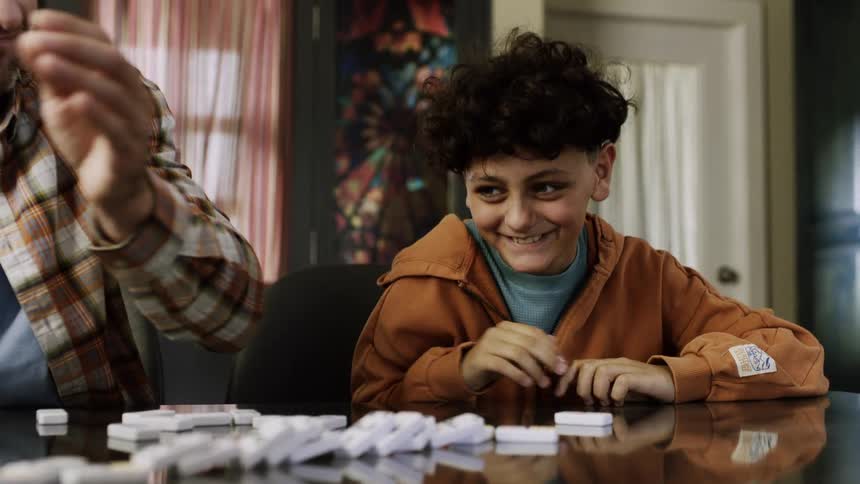 A smiling father and his son play a game of dominoes together at a table with the father playfully messing up the tiles at the end