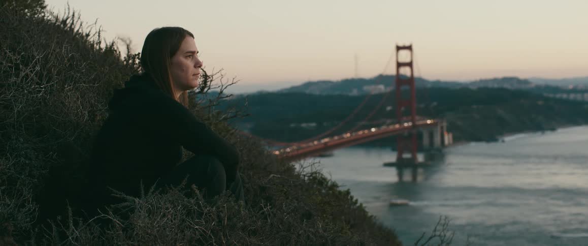 A woman sits on the hill with a large bridge in the background