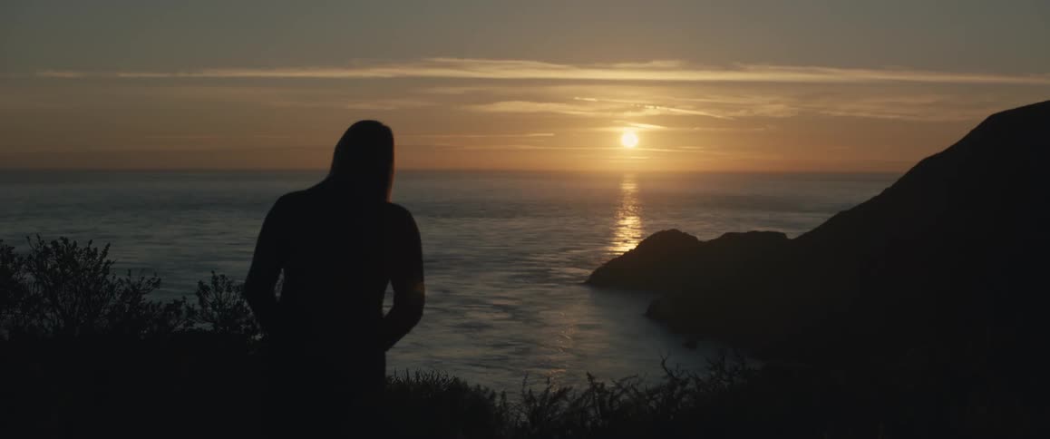 A person walks toward rocky cliffs in the distance silhouetted against a golden ocean sunset with sunlight glinting off the water