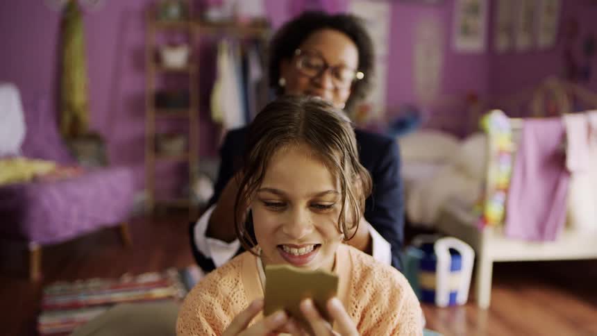 A woman helps a young girl with her hair in a cozy brightly colored bedroom