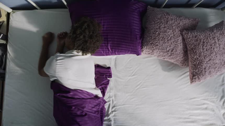 A woman lies in bed under a purple blanket wearing a white shirt begins to wake and stand up