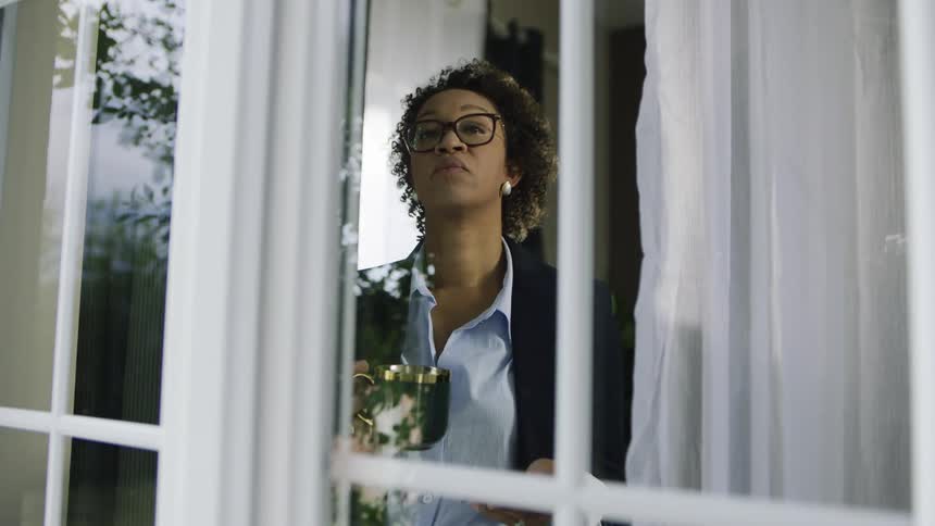 A woman drinks from a mug while checking her phone near a window