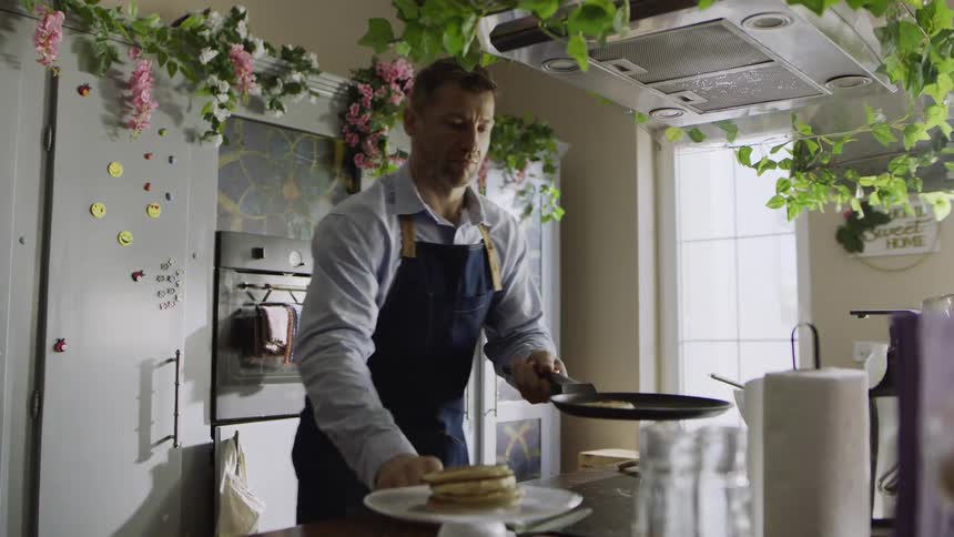 A man in an apron cooks and plates a stack of pancakes in a whimsical kitchen adorned with flowers and greenery