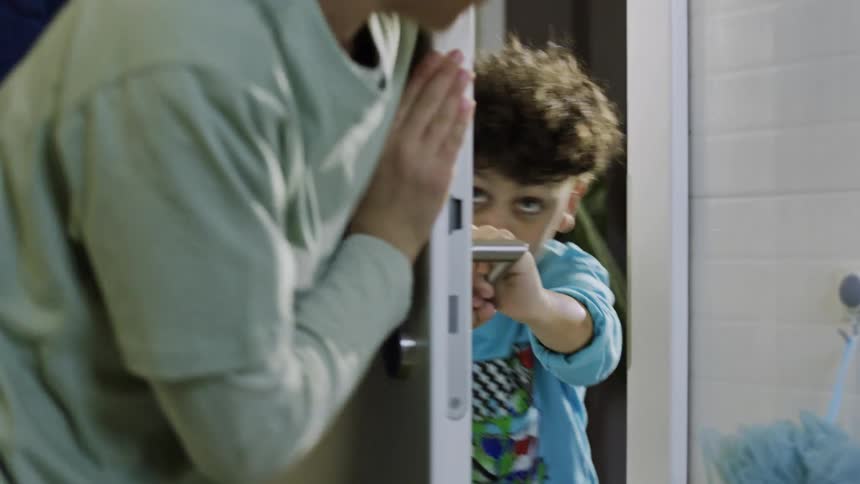 A teenage boy runs into the bathroom and plays around with his younger brother pushing the door back and forth before letting him enter