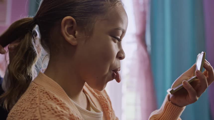 A smiling mother styles her daughters hair while the girl playfully makes faces at her reflection in a small hand mirror