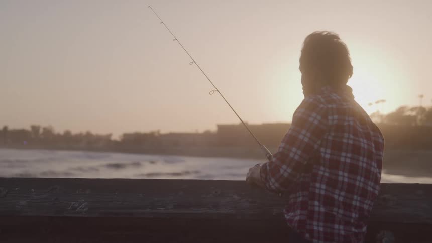 An older asian man fishes from a pier his silhouette framed against the warm glow of the sun over the ocean