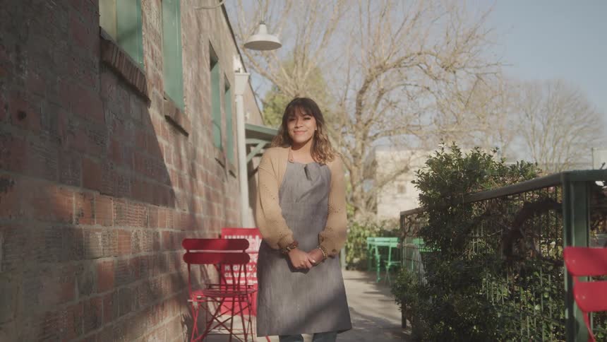 A young woman in an apron stands outside a caf smiling warmly in the sunlight