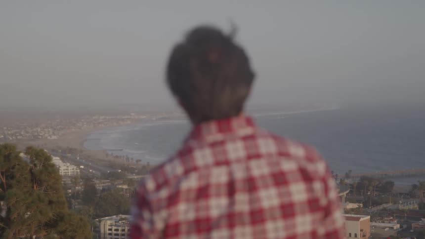 Man in a red plaid shirt overlooks a coastal city from a scenic high point