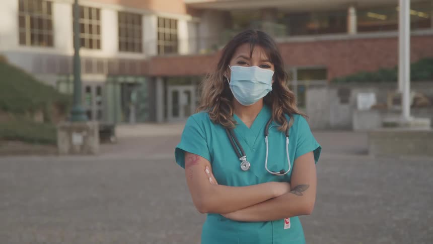 A female nurse wearing a face mask and teal colored scrubs stands with her arms crossed outside of a hospital building looking forwrd