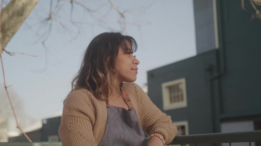 The face of a woman who is standing outdoors leaning her arm on a handrail looking to the left and she has a subtle smile on her face