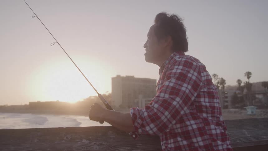 A man in a red plaid shirt stands on a pier holding a fishing rod and looking out at the ocean during sunset