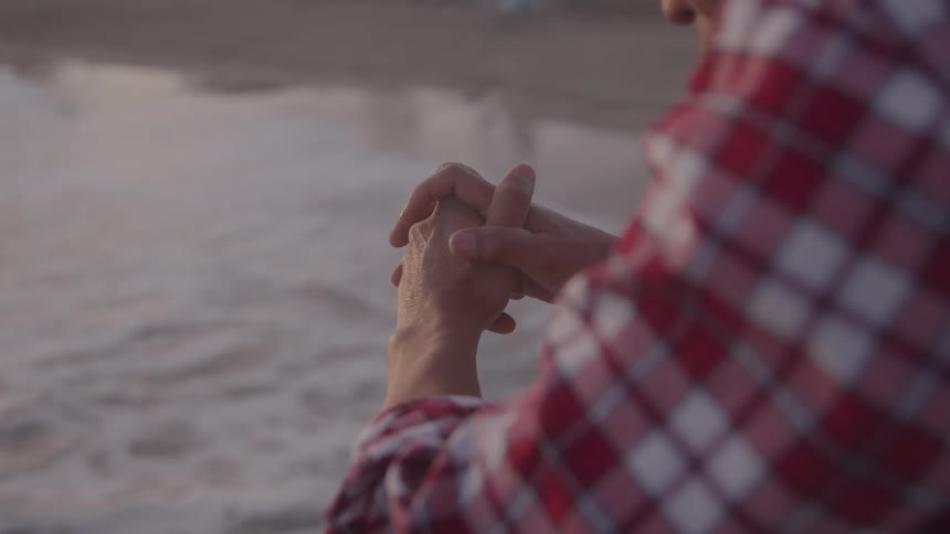 A man in a red and white checkered shirt stands near the shoreline clasping his own hands in a pensive meditative posture as waves roll in front