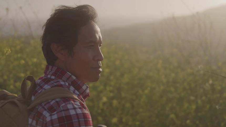 An older asian man in a red plaid shirt with a canvas backpack stops while hiking and gazes at the field of wildflowers with hazy hills and sun in the background
