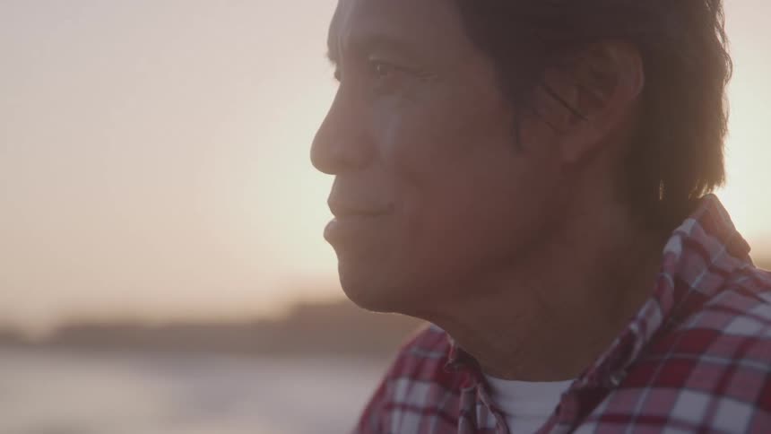 A man wearing a red and white plaid shirt stands near the ocean at sunset