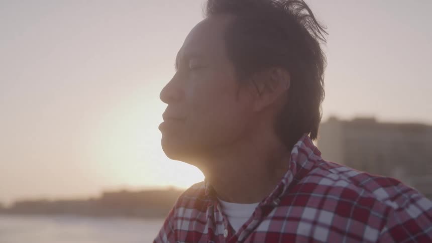 A closeup profile of an asian man with his eyes closed enjoying the warm sunlight while standing on a pier at sunrise or sunset