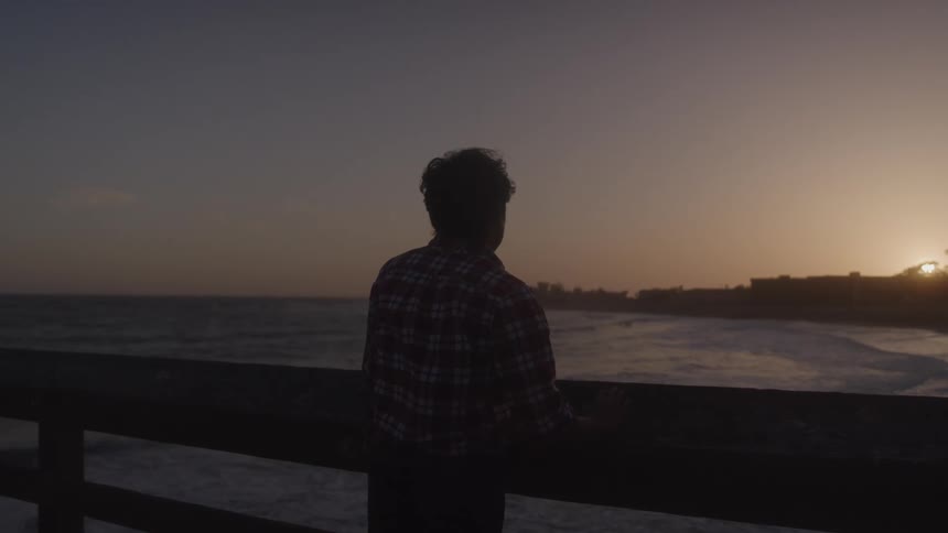 An older asian man in a red plaid shirt stands silhouetted on a pier gazing at the ocean as waves crash during sunset