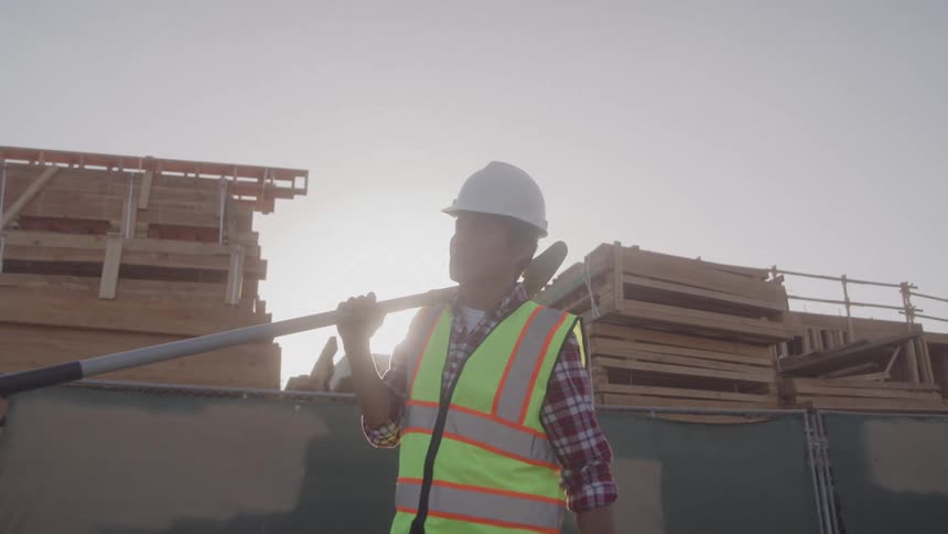 Construction worker in safety gear carries a shovel over shoulder at a building site