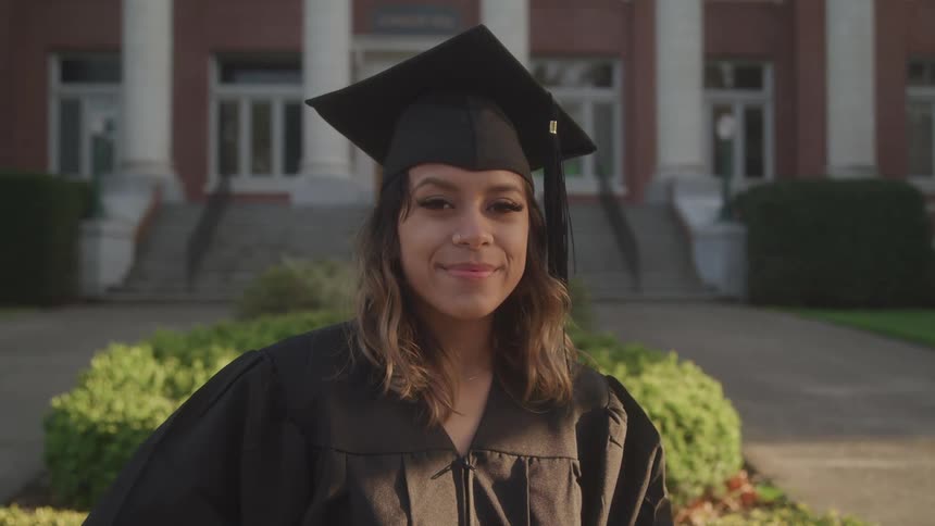 A young woman in a graduation cap and gown smiles confidently outside a university building