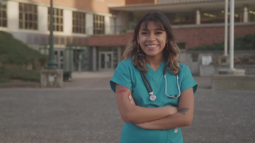 A smiling female healthcare worker stands confidently with arms crossed outside a building