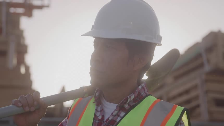A construction worker stands in sunlight holding a metal pole at a job site