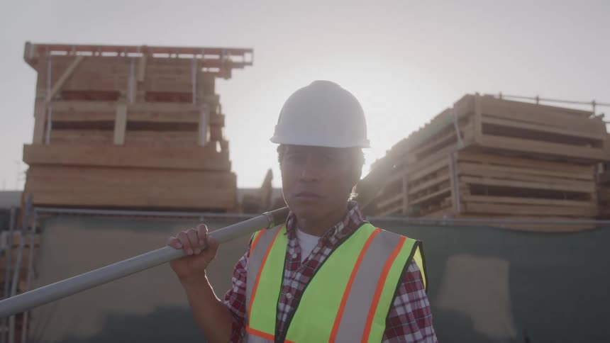 A male construction worker wearing a white hardhat and yellow vest holds a shovel over one shoulder while standing in front of a temporary fence with stacked pallets and a bright white sky in the background behind it