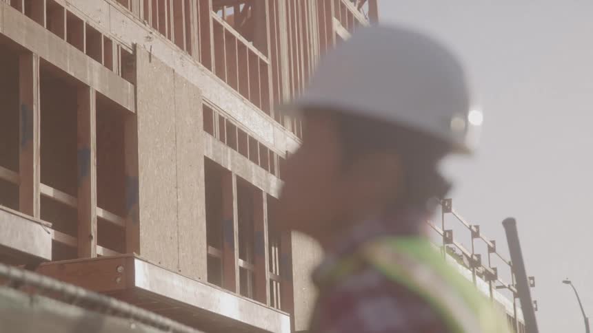 A construction worker wearing a hard hat and safety vest stands in soft focus in the foreground with the sunlit wooden frame of a building under construction sharp in the background