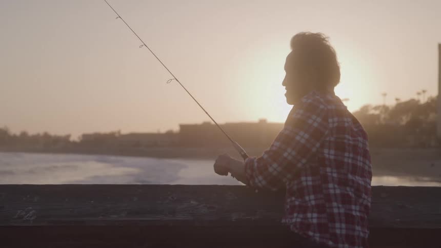 An asian man is silhouetted by the sun as he fishes with a rod and reel from a coastal pier at sunrise or sunset