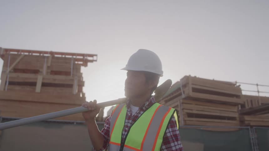 A construction worker wearing a green vest and a helmet holds a shovel outdoors at a construction site