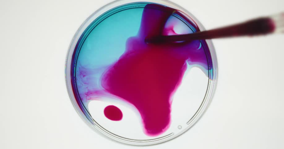 A pipette dispenses red liquid into a petri dish forming a spreading abstract pattern over blue liquid