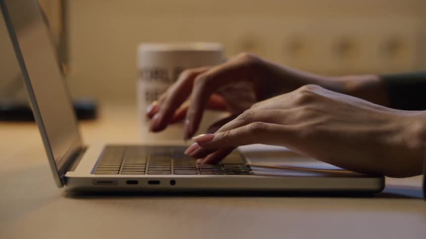 Hands type on a laptop keyboard beside a dark coffee mug on a wooden surface
