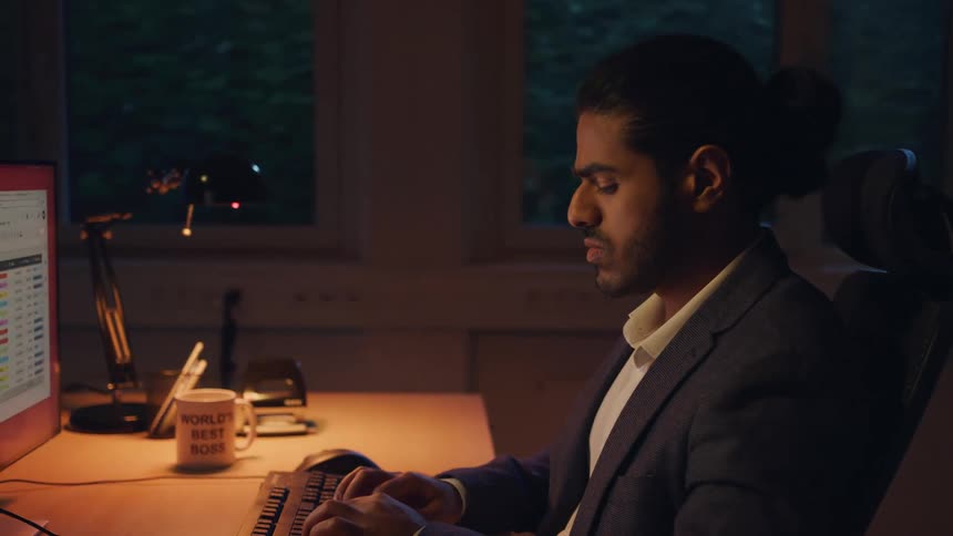 A man in a suit works intently on a computer at a dimly lit office desk