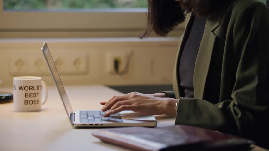A young woman typing on a laptop that is on a table in front of her and she picks up a mug to drink
