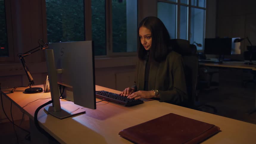 A woman works late at a desk in a dim office with large windows continuing her tasks by lamp light after someone turns off the overhead lights