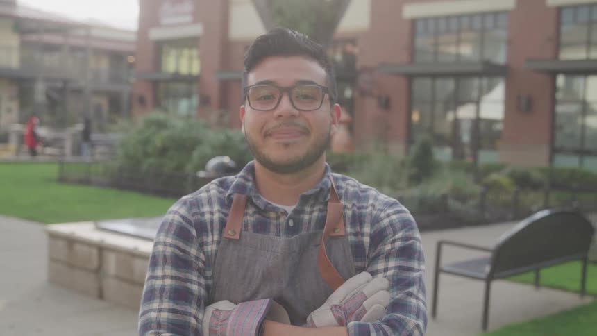 A young man wearing reading glasses and a jumpsuit stands with his arms crossed looking straight ahead with a subtle smile