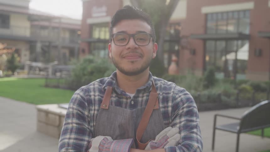 The face of a young man wearing reading glasses and has his arms crossed forward while having a subtle smile