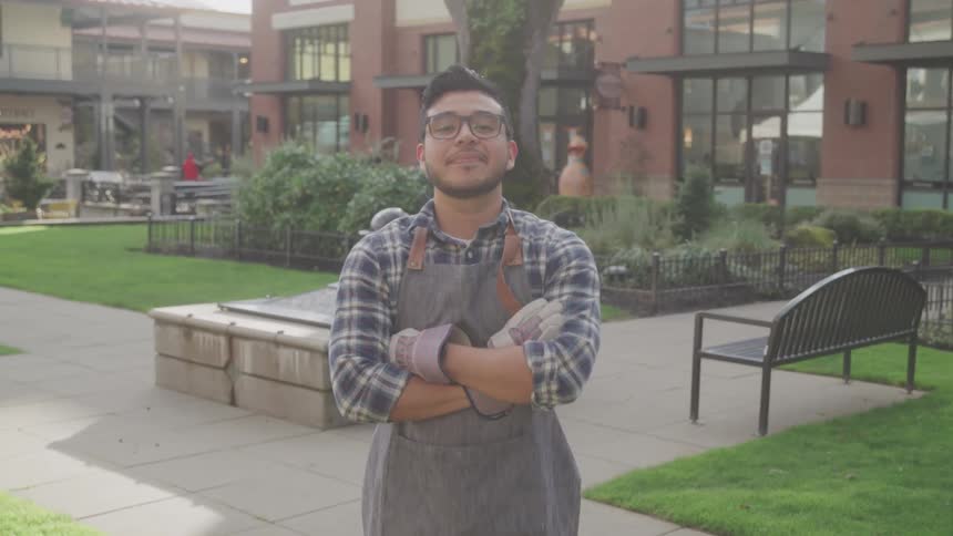 A smiling man in an apron stands confidently with folded arms in an urban courtyard garden