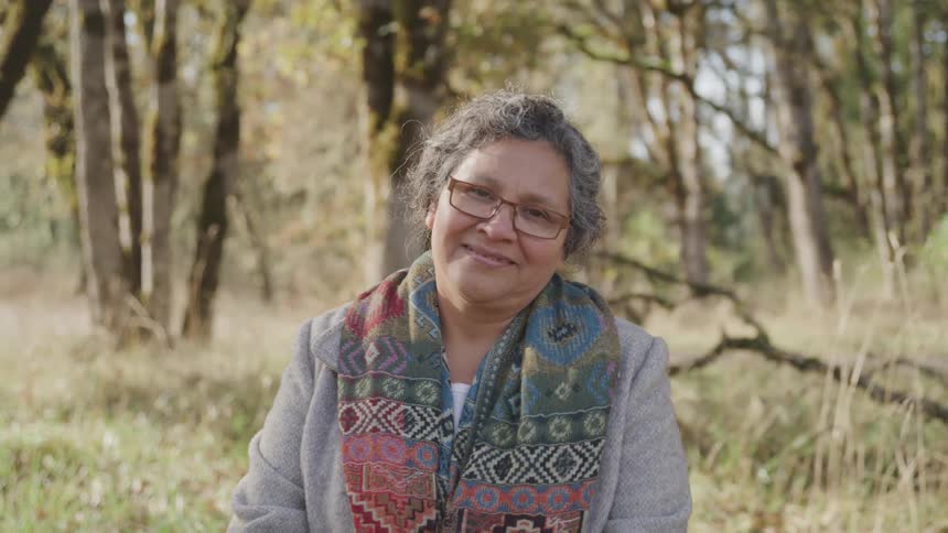 A smiling woman is seated outdoors in a sunlit forest facing the camera