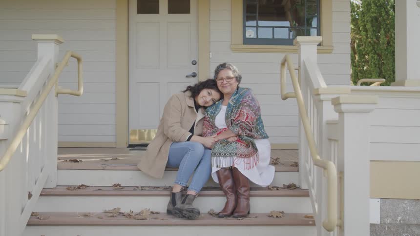 A granddaughter lovingly rests her head on her grandmothers shoulder as they sit on a porch step