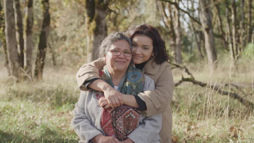 A young woman hugs her mother who is wearing glasses in a forest woodland around midday both are wearing winter jackets