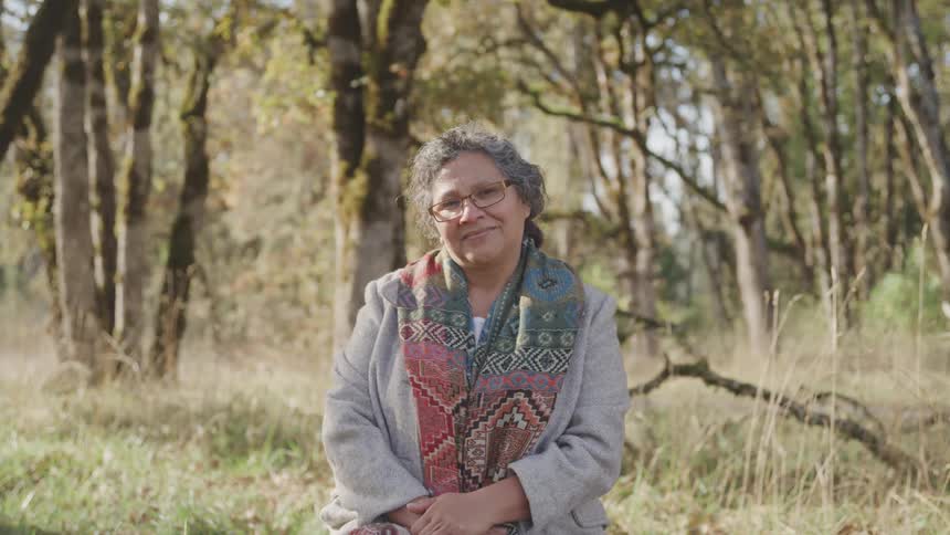 An older woman sits peacefully in a forest clearing smiling at the camera