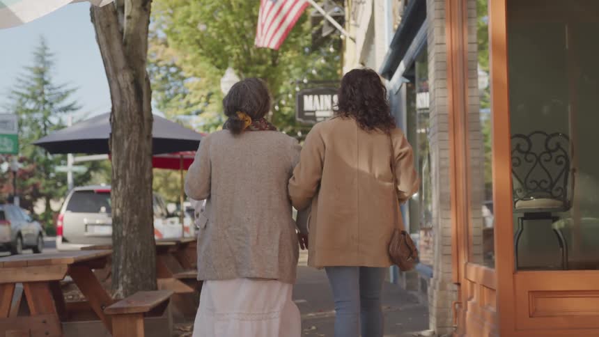 Two women walk arminarm down a sunny treelined sidewalk in a quaint town