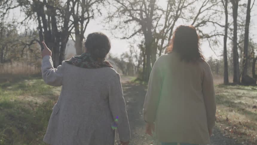 Two women walk side by side along a dirt path flanked by dry trees beneath clear skies