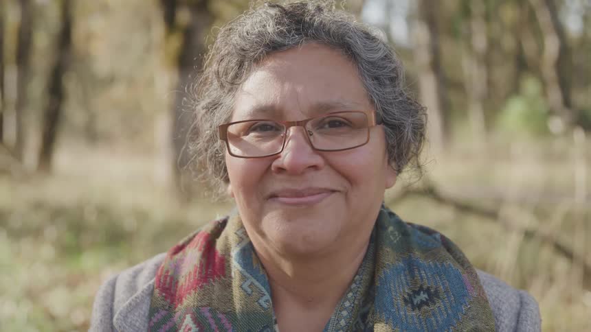 An elderly woman with a gentle smile outdoors near many trees