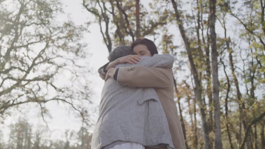 A younger woman and an older woman embrace tightly surrounded by tall trees with sparse leaves