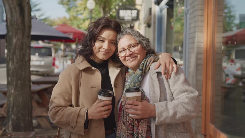 A young woman hugging her grayhaired mother and they are holding cups of coffee and wearing warm clothes