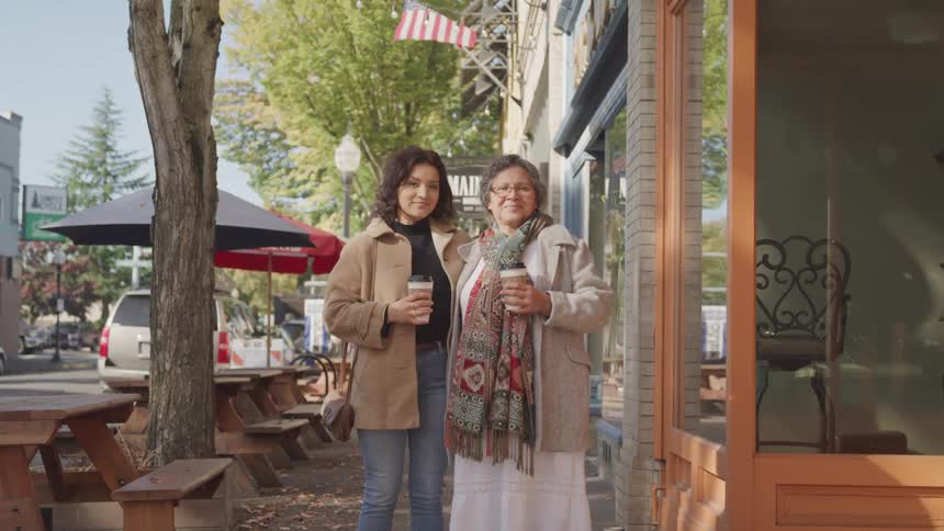 Younger and older female standing in front of a coffee shop holding coffee cups