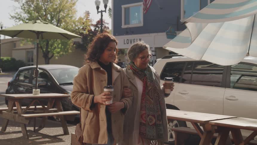 Two women walk along a street holding coffee cups one in a brown jacket and the other in a gray coat with a colorful scarf
